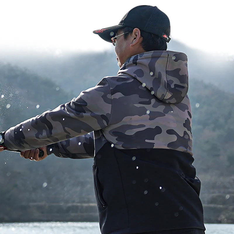 Person wearing a camouflage jacket and cap, fishing by a lake with mountains in the background.